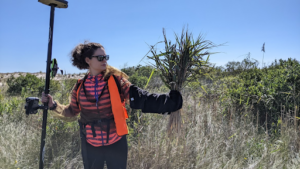 Alexandra Evans stands in a marsh, holding equipment in one hand and looking at grasses held in the other hand